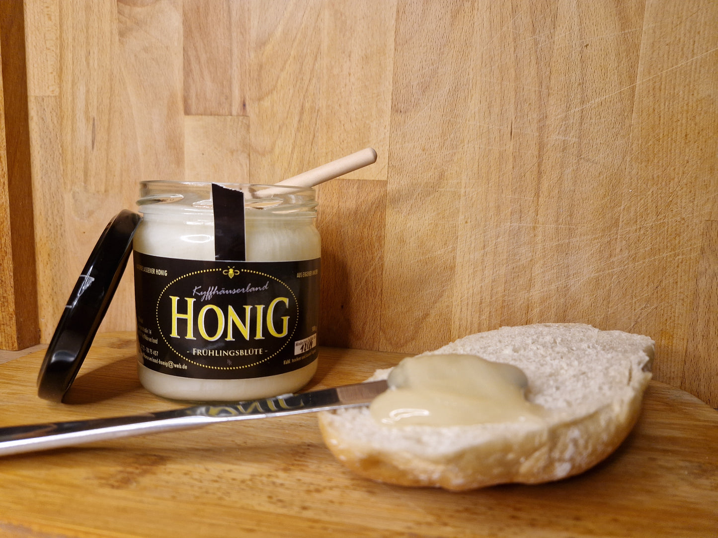 Ansicht eines feincremigen Frühlingsblütenhonigs im Glas. Der feincremige Honig verläuft au einem Brötchen.